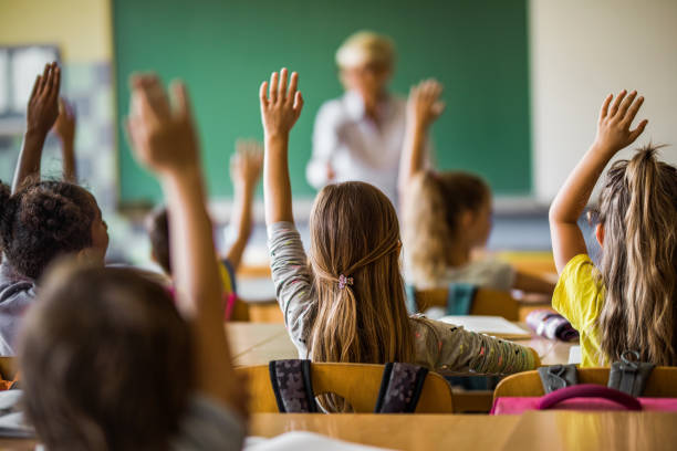 Rear view of large group of students raising their arms to answer the question on a class at elementary school.